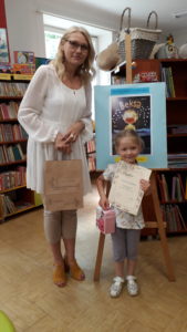 W bibliotece dziewczynka odbiera nagrodę od bibliotekarki, w tle sztaluga z pracą konkursową.
